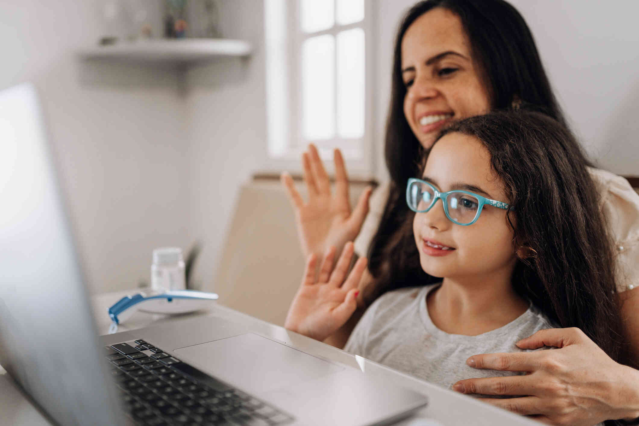 A young girl wearing glasses sits on a woman's lap and the two of them smile and wave towards a laptop screen in front of them.